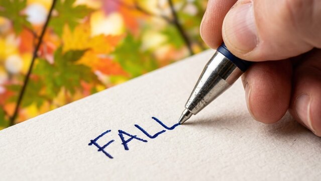 Person writing the word Fall on paper with autumn leaves background