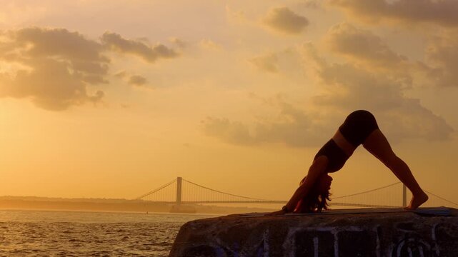 Silhouette of woman doing downward dog pose at sunset, New York. Angle of yoga pose on concrete ledge with suspension bridge across water under yellow sky.