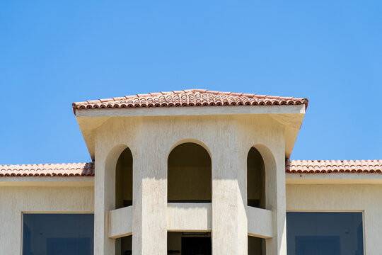 Beautiful architectural detail of Mediterranean style villa featuring beige stucco facade, arched openings, and terracotta tiled roof against clear bright blue sky.