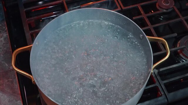 Boiling water bubbling in a gray pot on a gas stove. Steam rises from the hot liquid as rolling bubbles cover the liquid surface during cooking.