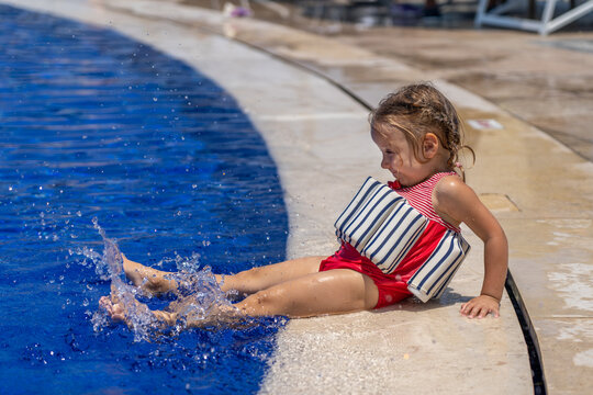 Adorable Young Girl Wearing Striped Puddle Jumper Floating Vest Happily Splashing Her Feet in Sunny Outdoor Swimming Pool During Fun Summer Vacation Day