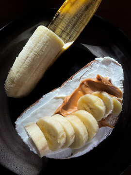 Top view of healthy breakfast toast spread with thick Greek yogurt, creamy peanut butter, and fresh sliced bananas served with a half-peeled banana on a dark plate.
