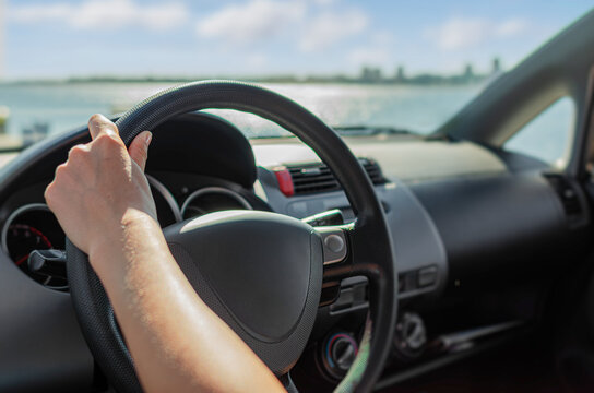A person holds the steering wheel while driving a car along a scenic coastal road under a sunny sky.