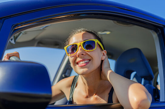 A happy young woman wearing bright yellow sunglasses smiles while driving a blue car on a sunny day.