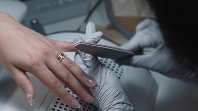 Manicure technician shapes acrylic nail using salon file during professional cosmetic procedure in beauty salon