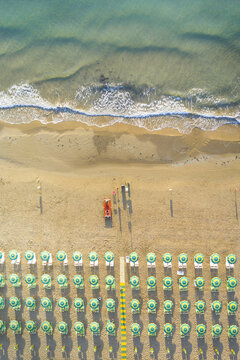 Aerial view of rows of green and white beach umbrellas on a sandy shore with turquoise sea waves lapping the coastline in Apulia, Italy.