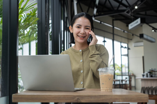 Professional woman working on a laptop in a modern cafe while talking on a smartphone, representing remote work, business communication, productivity, freelancing, and flexible career lifestyle.