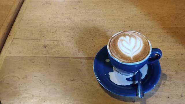 Blue cup holds creamy coffee art. Heart design floats atop steamed milk. Warm wooden table adds rustic charm. Simple, inviting drink perfect for quiet moments. Cafe setting feels cozy and relaxed