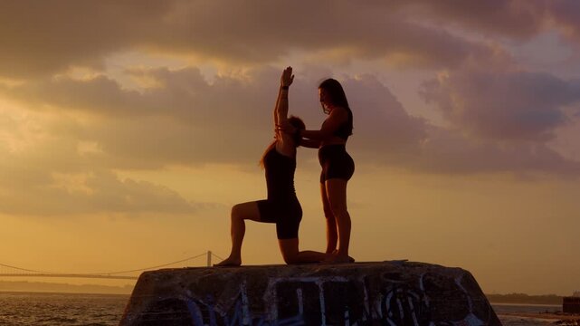Instructor assisting student in yoga pose near bridge, New York. Kneeling stretch on stone structure against golden sunset sky and suspension bridge.