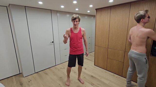 Two men perform acrobatic lift indoors. One man supports the other overhead. They smile during playful movement. Wooden walls and modern lighting frame scene. Floor is polished light wood