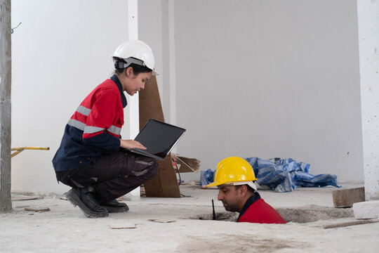 Construction engineers inspecting an underground utility access with a laptop during structural building assessment, highlighting foundation inspection, workplace safety, infrastructure maintenance.