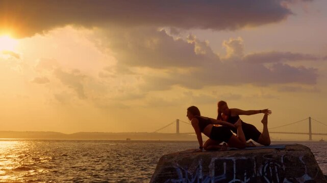 Quad stretch yoga posture on waterfront pier, New York, USA. Two female silhouettes performing mermaid pose at golden hour with distant bridge.