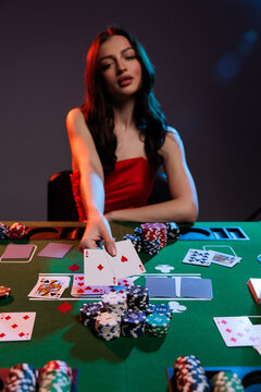 Beautiful young woman sitting at poker table with playing cards, chips and glass of champagne on dark green background
