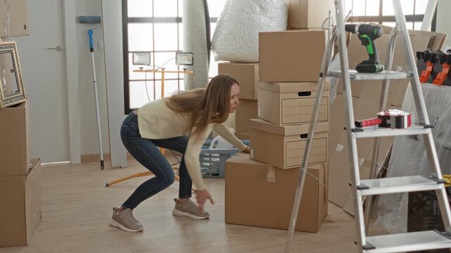 Woman holding lower back for backache while reaching stacked cardboard boxes and moving supplies in a building; frustration.