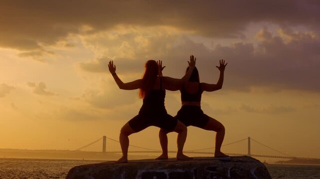Goddess pose yoga practice near ocean bridge, New York, USA. Two female silhouettes squatting on concrete block at sunset.