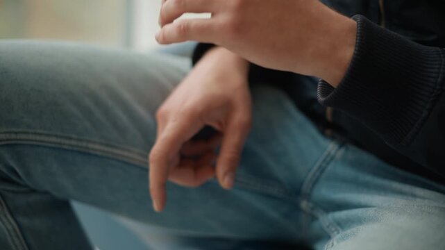 Close up of man hands resting on his lap with fingers interlaced wearing blue jeans and dark jacket. Soft lighting highlights skin texture details while background remains blurred focusing