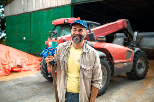 Smiling bearded farmer holding shovel by tractor at barn