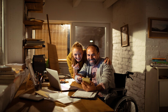 Man in wheelchair and woman using smartphone in home office at night