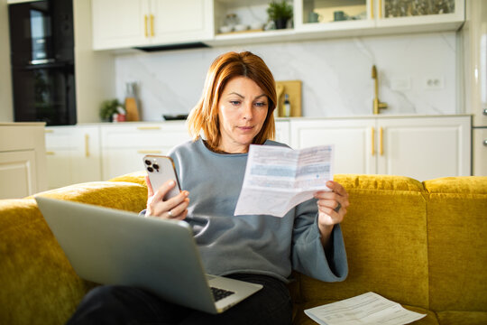 Woman reviewing household bills with phone and laptop on home sofa
