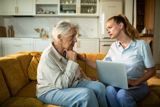 Caregiver supporting senior woman at home during laptop consultation