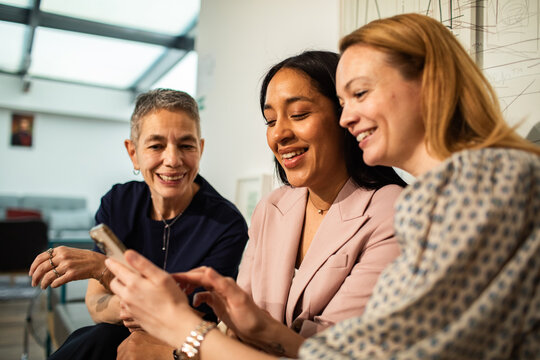 Three female colleagues sharing smartphone in modern office