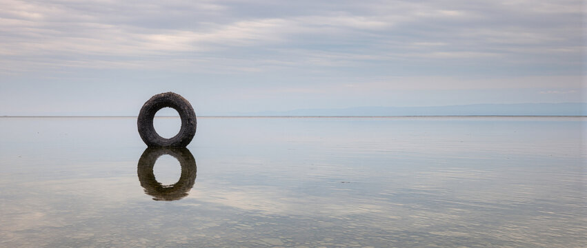 Large dark circular object standing in shallow still water under an overcast sky. Minimalist landscape with perfect symmetrical reflection
