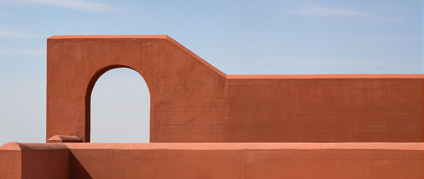Terracotta architectural structure with an archway and stepped wall against a clear blue sky. Minimalist geometric building exterior in bright sunlight