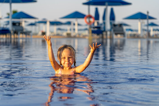 Happy young girl with arms raised splashing and playing in sunny resort swimming pool with blue umbrellas in background during fun summer vacation holiday trip