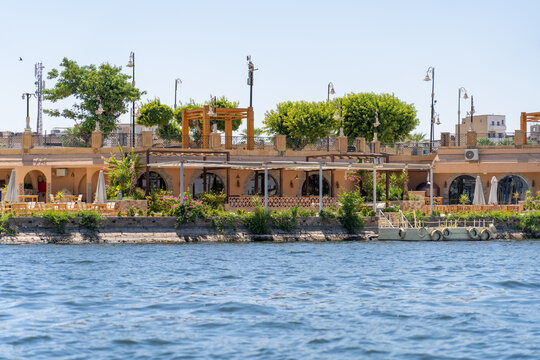 Scenic waterfront cafe terrace featuring traditional architecture, lush greenery, and outdoor dining tables situated along calm river under bright clear blue summer sky in Egypt