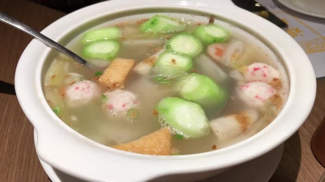 A top angle shot of a hot bowl of savory broth soup containing meatballs, tofu, and sliced zucchini on a table.