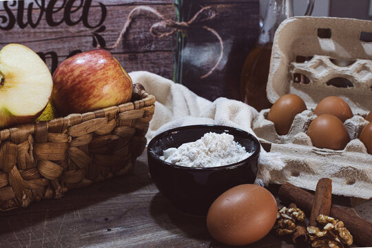 Assorted organic baking ingredients, including fresh apples, eggs, flour, cinnamon, and walnuts, artfully arranged on a weathered wooden surface, preparing for a homemade recipe