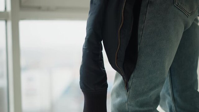 Close up of person wearing blue jeans and holding bottom hem of dark jacket with visible stitching and zipper details. background shows out of focus window suggesting indoor setting