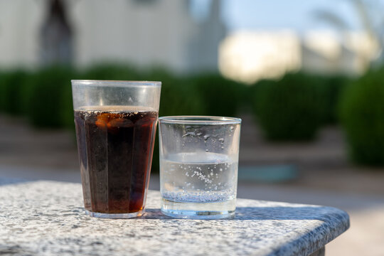 Refreshing pair of glasses featuring chilled dark fizzy cola beverage alongside clear sparkling carbonated water glass placed on outdoor textured marble table in daylight