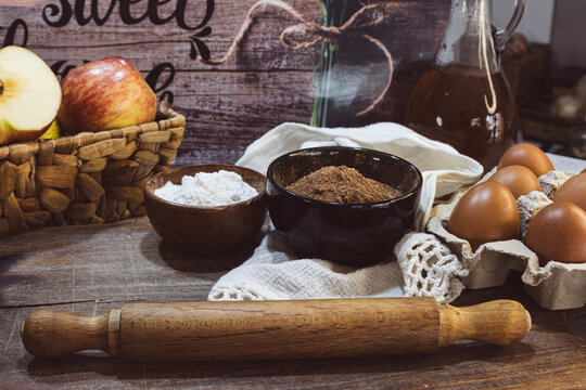 Baking ingredients on a rustic wooden table, cocoa powder, flour in a bowl, rolling pin and fresh organic eggs in a carton for homemade preparation.
