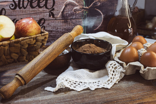 Cocoa pwder in a black bowl, a wooden rolling pin, a white cloth, and brown eggs in an egg carton arranged on a rustic wooden surface, representing home baking and fresh ingredients.