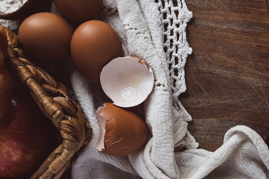 Fresh organic apples and brown eggs, including a cracked shell on a textured linen cloth on a distressed wooden surface, preparing for baking or healthy meal preparation.