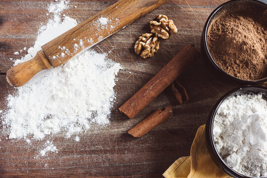 Baking ingredients on rustic wooden table displaying flour, cocoa powder, walnuts, and cinnamon sticks with a rolling pin, preparing for home cooking and making desserts