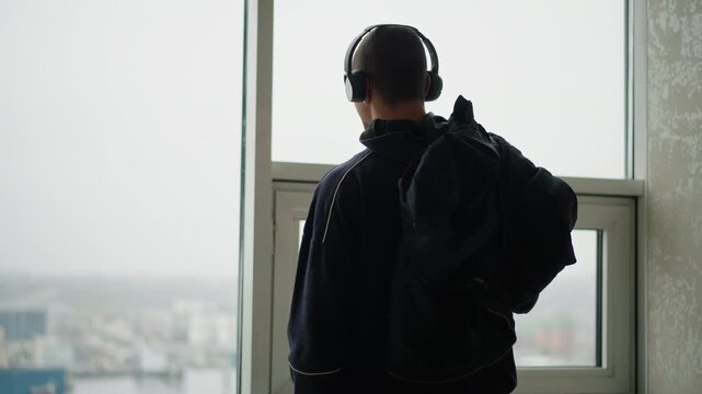 Man wearing headphones and carrying backpack stands still by large window facing cityscape. Silhouette shows consistent posture with slight head movement. Soft natural light illuminates