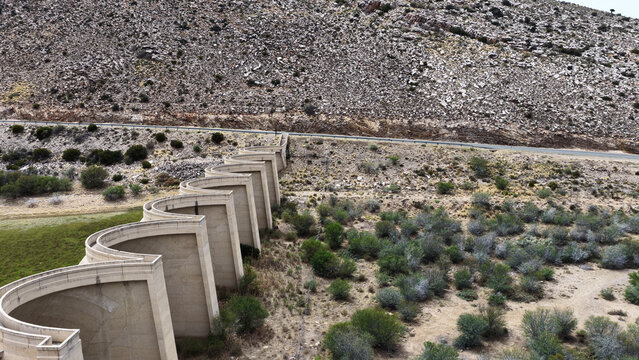 Aerial view of the serpentine concrete dam wall in the Little Karoo, surrounded by arid rocky hills and sparse vegetation in Garden Route District Municipality, Western Cape, South Africa.