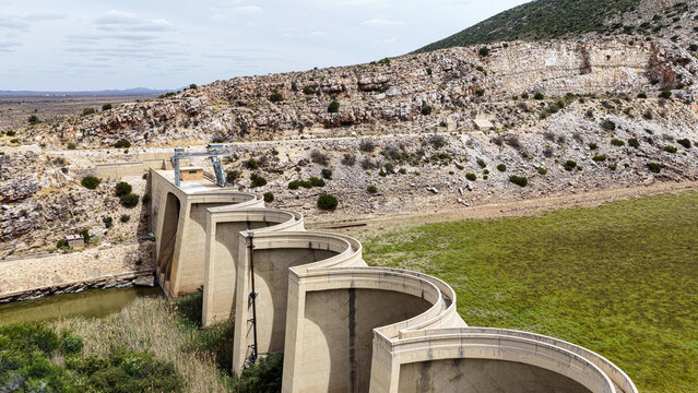 Aerial view of the curved concrete wall of the dam in the Little Karoo, surrounded by rocky hills and arid landscape in Garden Route District Municipality, Western Cape, South Africa.