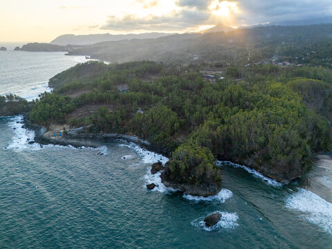 Aerial view of lush green cliffs and forest meeting the ocean waves along the rugged coastline at sunset in Pacitan, East Java, Indonesia.