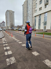 Person rides unicycle on city street. Wearing helmet and backpack. High-rise buildings line the background. Pedestrian crosswalk nearby. Overcast sky above the scene © Pavel Losevsky