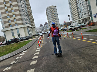 Man rides unicycle on city street. Buildings loom behind him in background. Orange cones line the road ahead. Grey sky overhead. Urban setting with parked cars. Pedestrian path beside asphalt © Pavel Losevsky