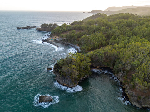 Aerial view of the rugged coastline with lush green forest, rocky cliffs, and ocean waves crashing against the shore in Pacitan, East Java, Indonesia.