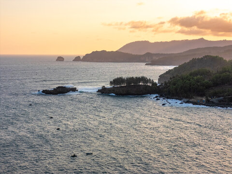 Aerial view of the coastal landscape in Pacitan with small fishing boats on the water, rocky cliffs, and lush vegetation under a warm sunset sky in Pacitan, East Java, Indonesia.