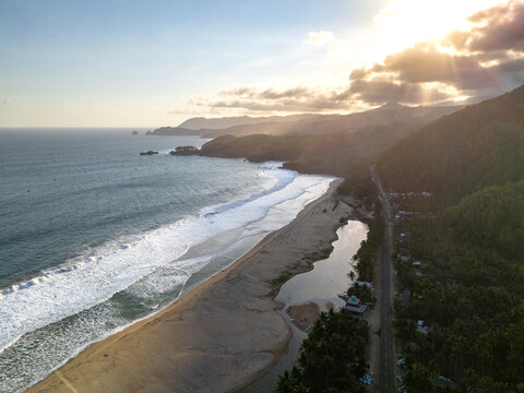 Aerial view of the coastline with waves crashing on the sandy beach, a coastal road alongside lush vegetation, and the warm glow of the setting sun in Pacitan, East Java, Indonesia.