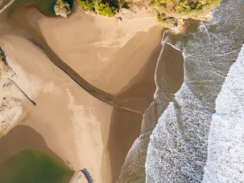 Aerial view of a sandy beach where a river meets the ocean, with white waves crashing on the shore and lush green vegetation along the coastline in Pacitan, East Java, Indonesia.