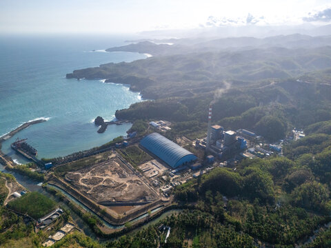 Aerial view of the coal-fired power plant with a tall chimney and large blue warehouse located along the lush green coastline of the Indian Ocean in Pacitan, East Java, Indonesia.
