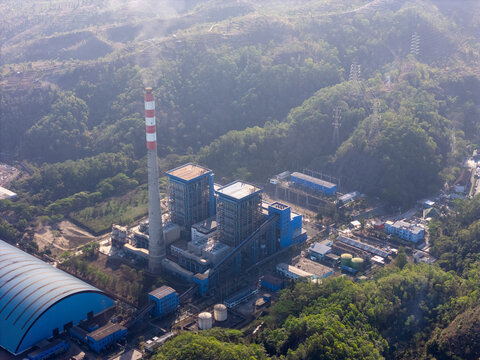 Aerial view of a power plant with a tall chimney and industrial buildings nestled among lush green hills in Pacitan, East Java, Indonesia.