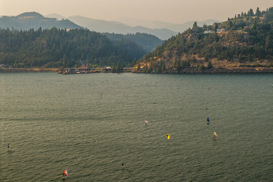 Aerial view of windsurfers and kitesurfers on the Columbia River under hazy skies from wildfire smoke, with forested mountains in the background in Hood River, Oregon, United States.